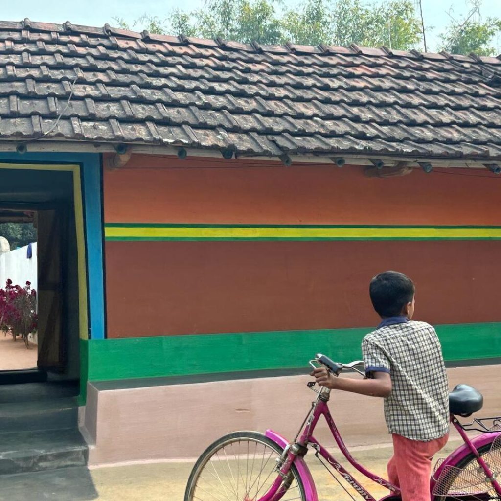 Un garçon tenant un vélo rose devant une maison colorée avec un toit en tuiles, illustrant une scène rurale paisible.